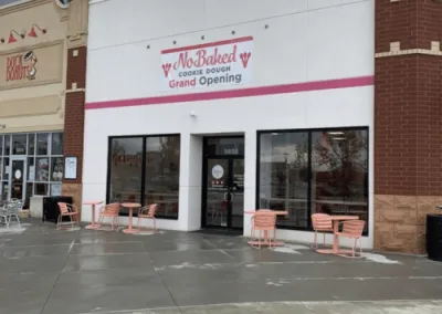 Exterior view of No-Baked Cookie Dough storefront with 'Grand Opening' signage, featuring a modern design with pink accents and outdoor seating, developed by Drake Architecture.