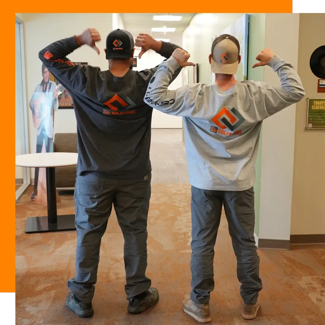 Two Cu Solutions technicians in branded long sleeve shirts and hats pointing to the company logo inside a Colorado office hallway