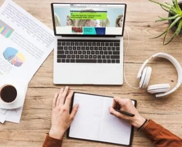 Remote worker taking notes beside a laptop, coffee, and headphones on a wooden desk.