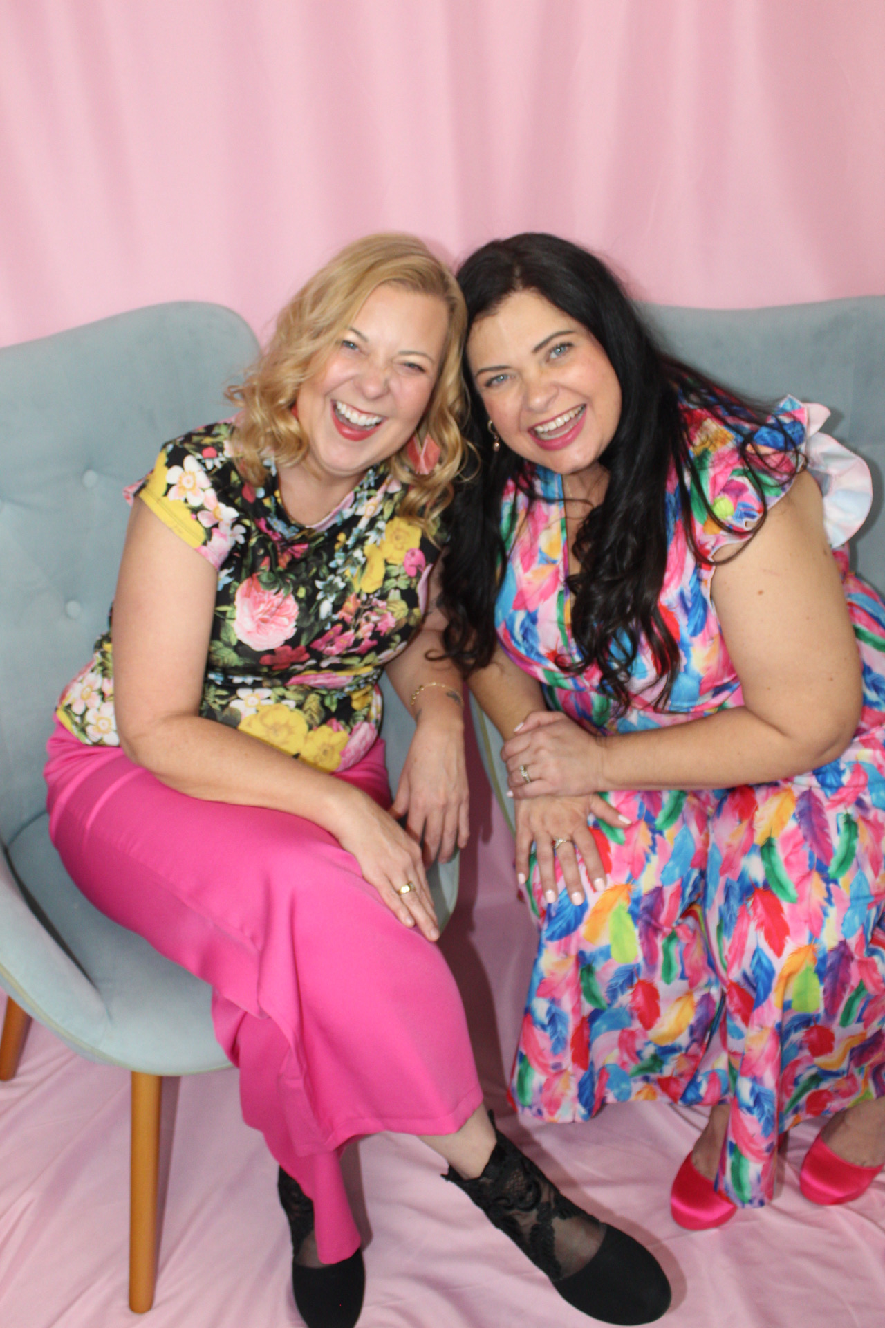 A tall brunette woman in a navy dress and a blonde woman in a pink jumpsuit stand back to back laughing with joy.
