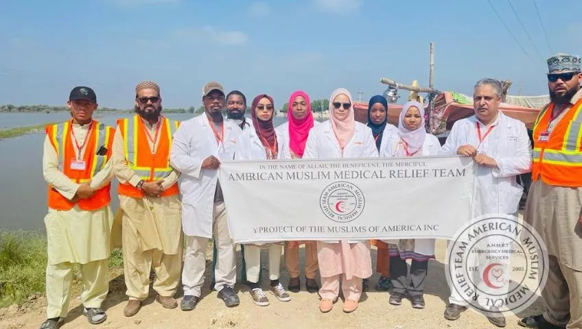 Members of the American Muslim Medical Relief Team (AMMRT) pose outdoors holding a banner during flood relief efforts in Pakistan. The team, consisting of medical professionals in white coats and safety vests, stands in front of a body of water, likely representing the flood-affected area they are serving.