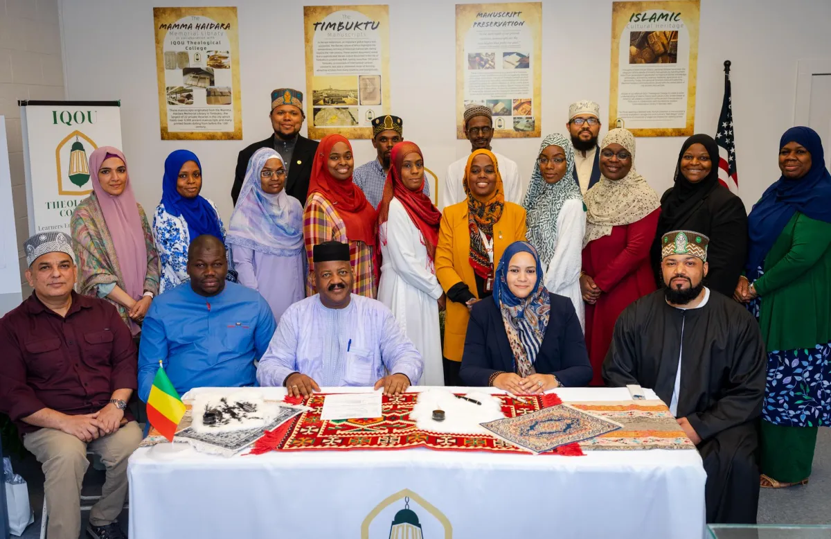 A diverse group of individuals, including faculty and guests, gather around a table at the IQOU Theological College for the signing of a memorandum with the Mamma Haidara Library. The table displays cultural artifacts and documents, while educational posters about Timbuktu, manuscript preservation, and Islamic history adorn the wall behind them.