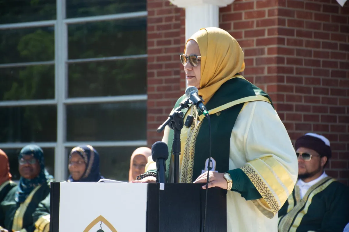 Sheikha Syeda Zainab Adams Gillani, wearing a yellow hijab, sunglasses, and a green and white robe with gold trim, delivers an address at a podium during the IQOU Theological College graduation. Special guests and teachers in traditional Islamic attire are visible in the background against a brick building facade.