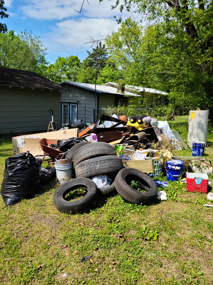 Open dump-style trailer with solid sides loaded with household items, attached to a pickup truck at a residential home in Northwest Arkansas