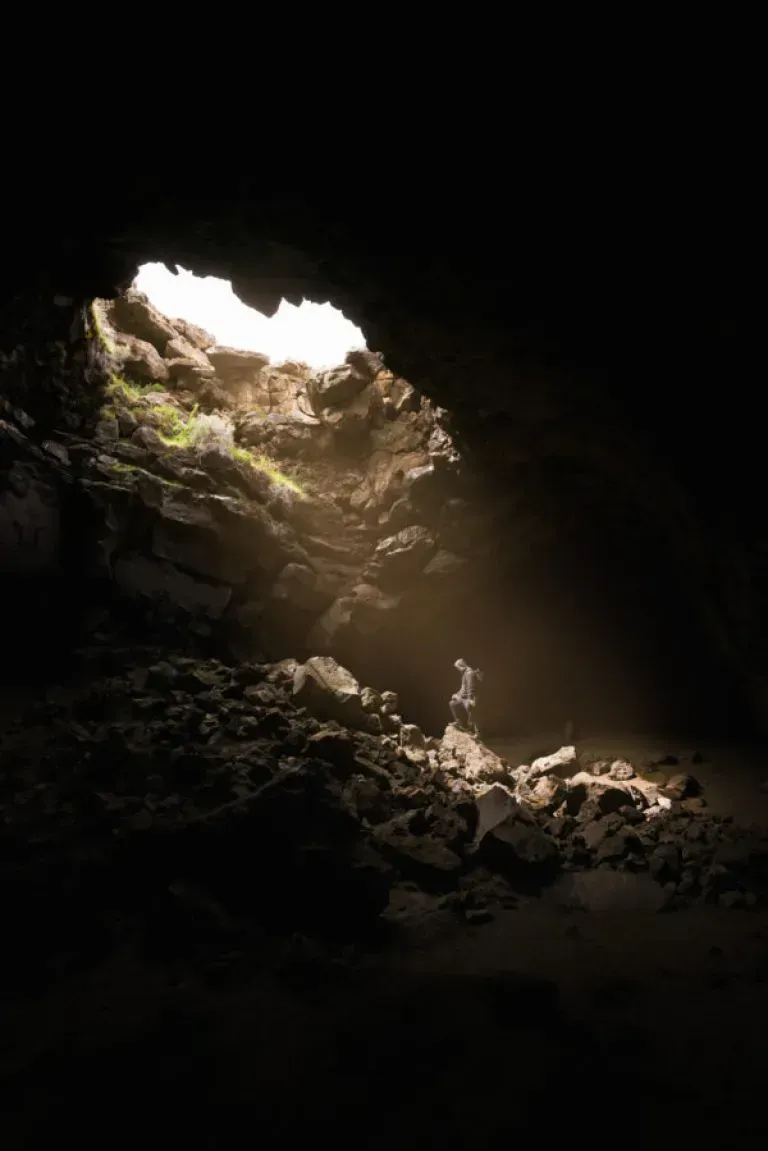 A person standing on a rocky surface inside a cave, with light streaming through the cave's opening, illuminating the rocks and creating a dramatic contrast between light and shadow.