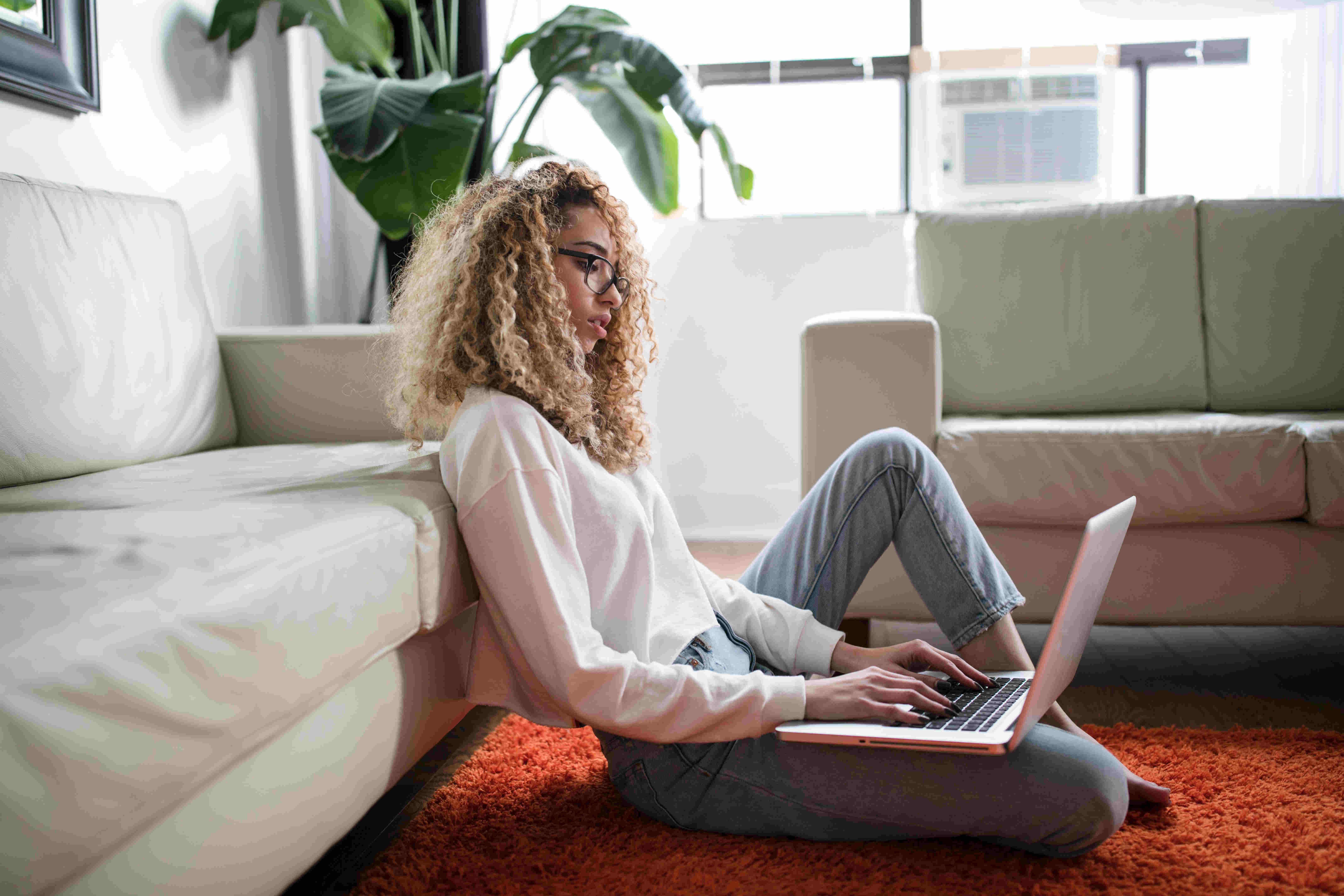 A person with curly hair, wearing glasses, sitting on the floor in a cozy living room, typing on a laptop. The scene includes a white sofa, indoor plants, and soft natural lighting.