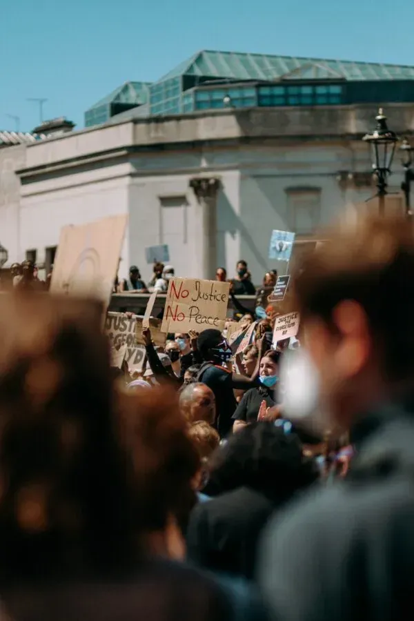 A crowd of protesters holding signs, including 'No Justice No Peace,' during a demonstration. The scene captures a diverse group of individuals in front of a building, with some wearing masks.