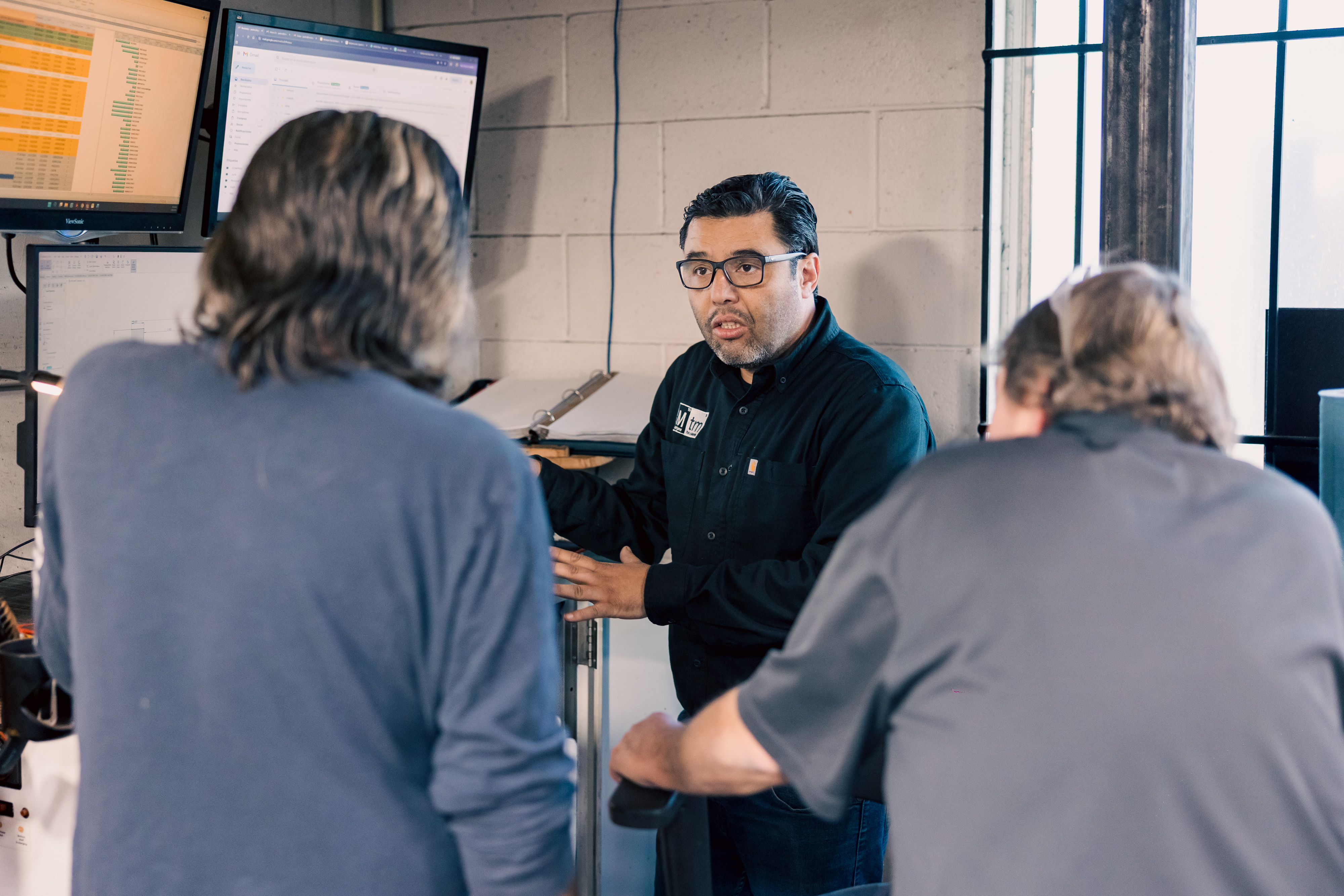Three people in an industrial office/workshop area discussing work in front of multiple computer monitors.