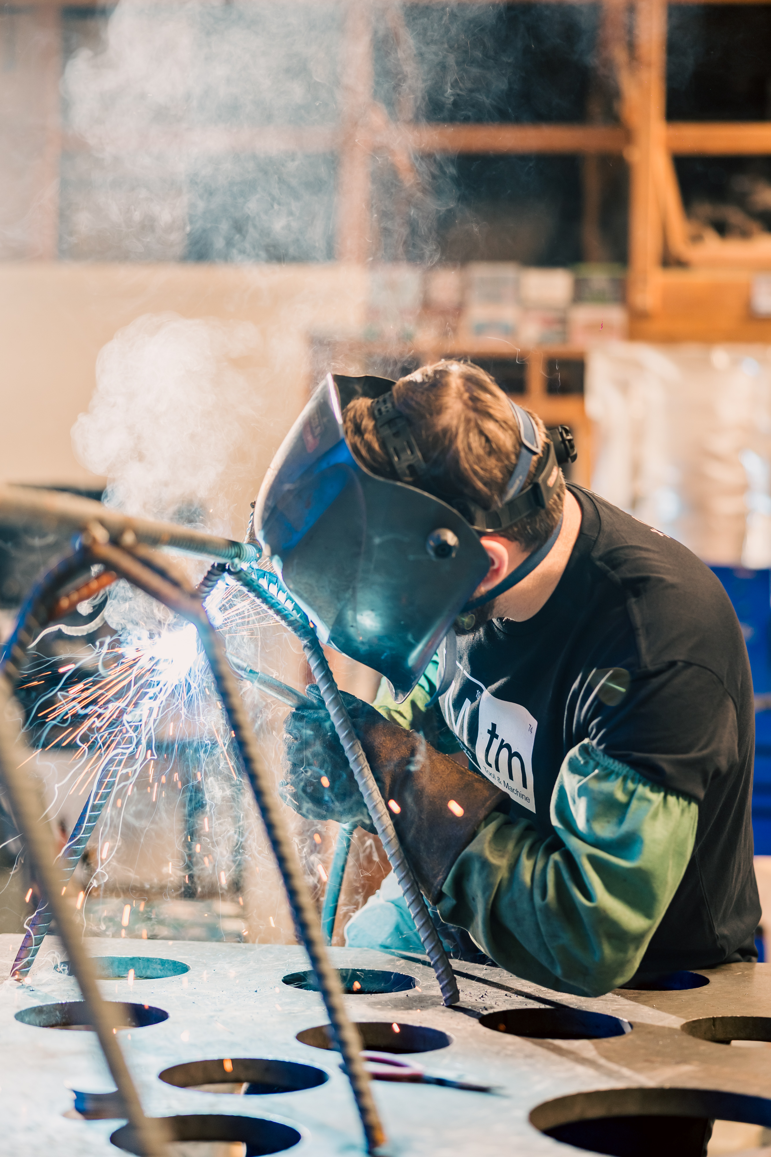 A fabricator welding on a perforated metal table, sparks and smoke in the workshop.