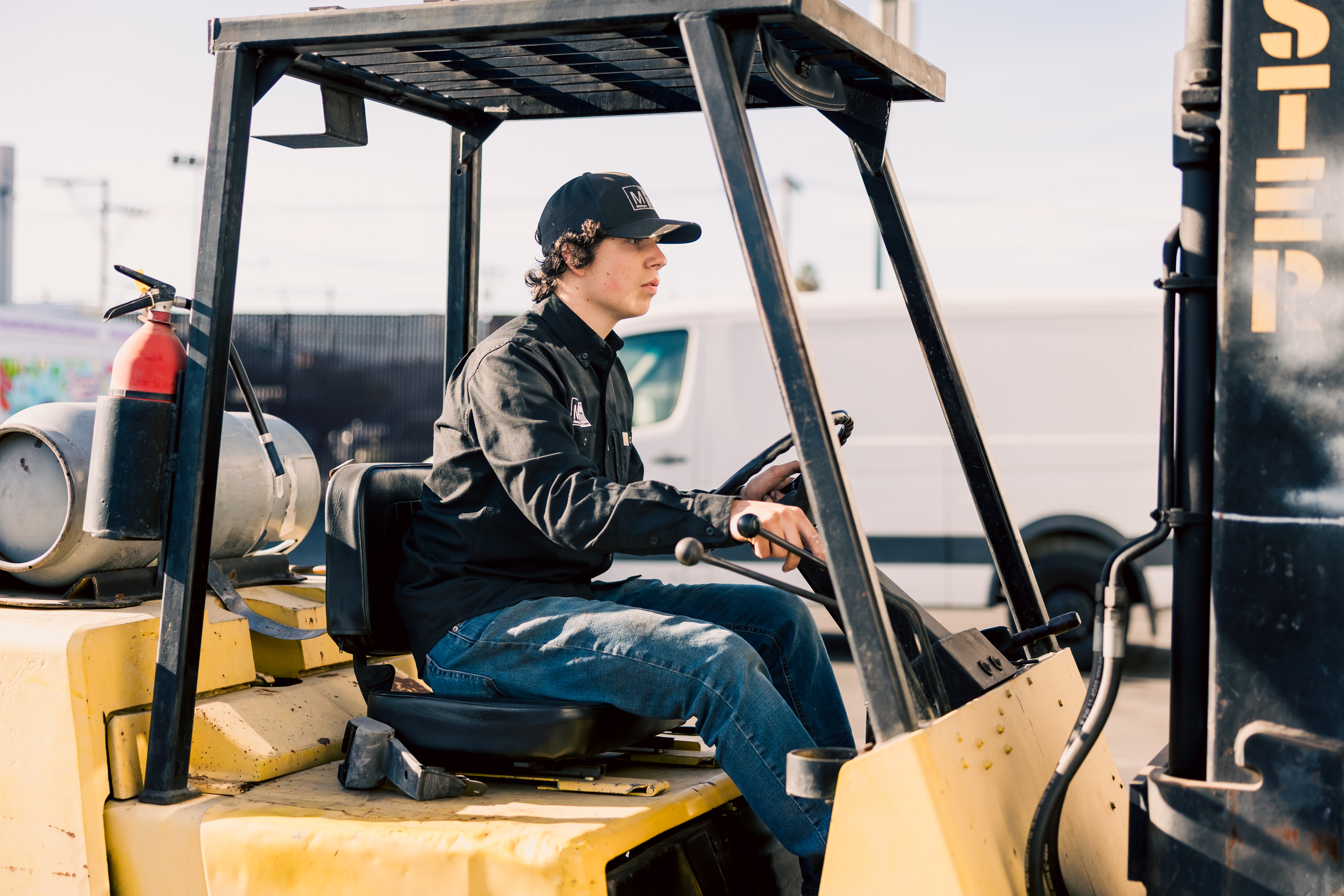 A man in a black cap and jacket operating a yellow forklift in an outdoor/loading dock area.