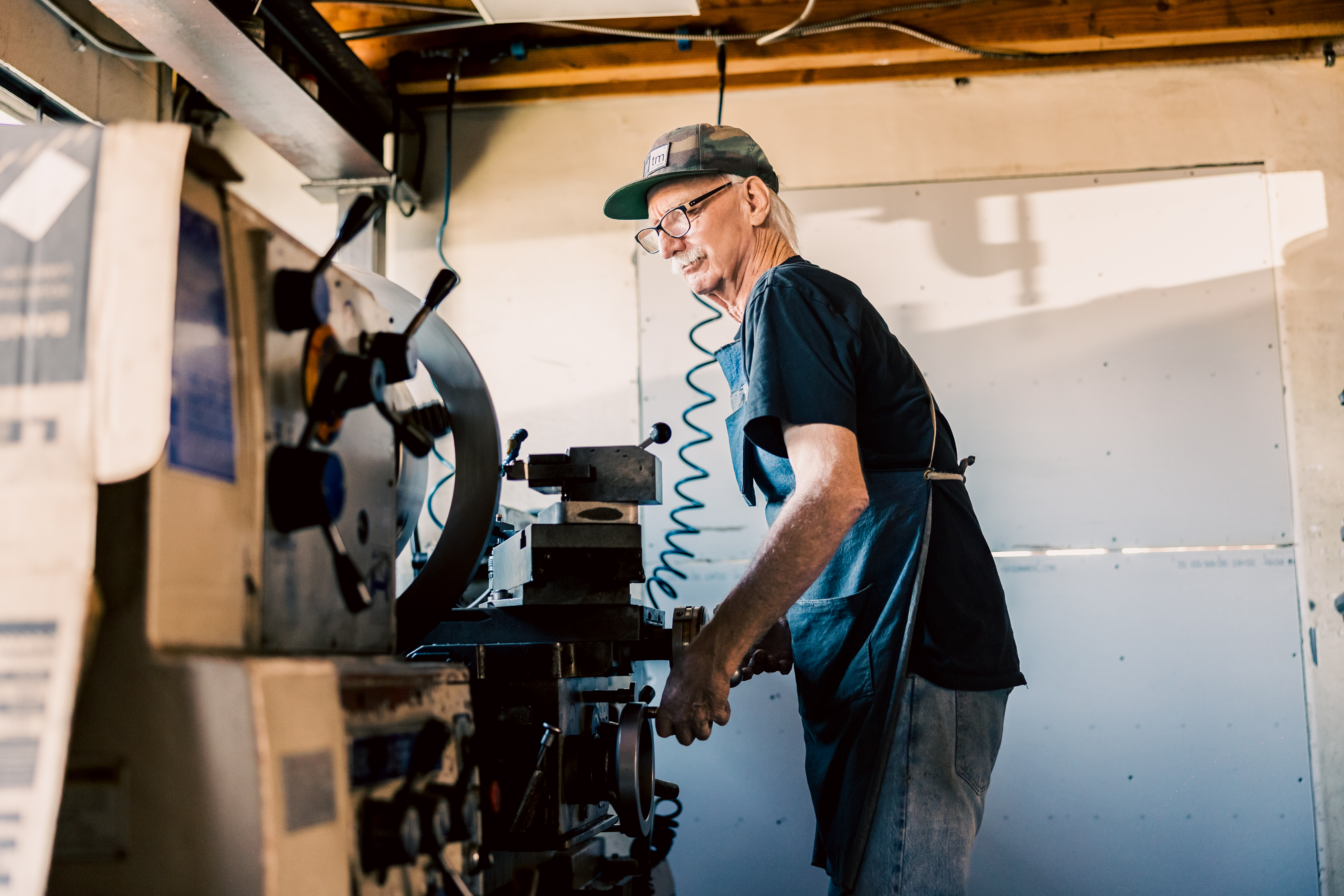 An older man with glasses and a cap is operating a large manual ACRA lathe, machining a large diameter metal ring in an industrial workshop.
