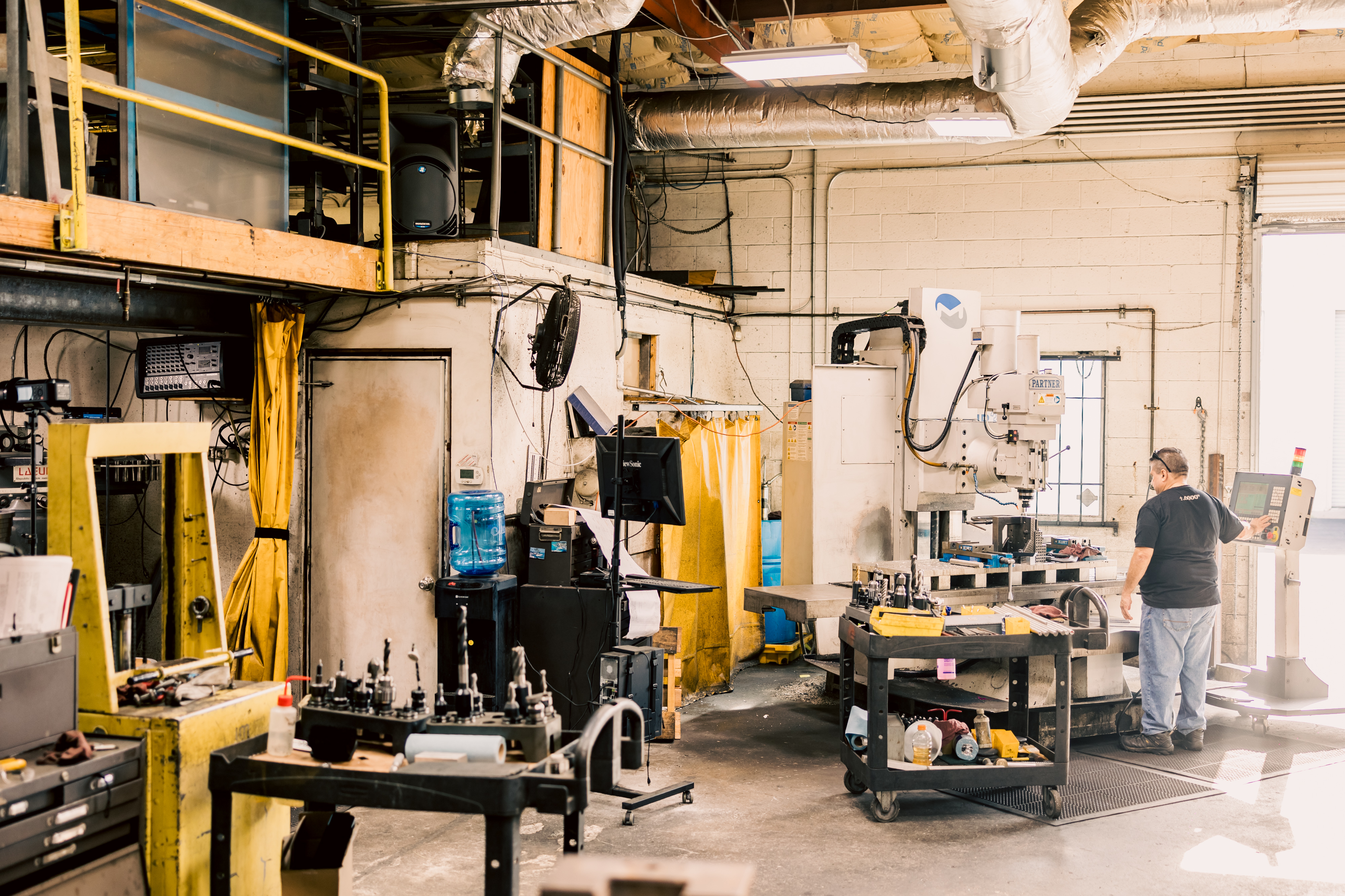 An older man with glasses and a cap is operating a large manual ACRA lathe, machining a large diameter metal ring in an industrial workshop.