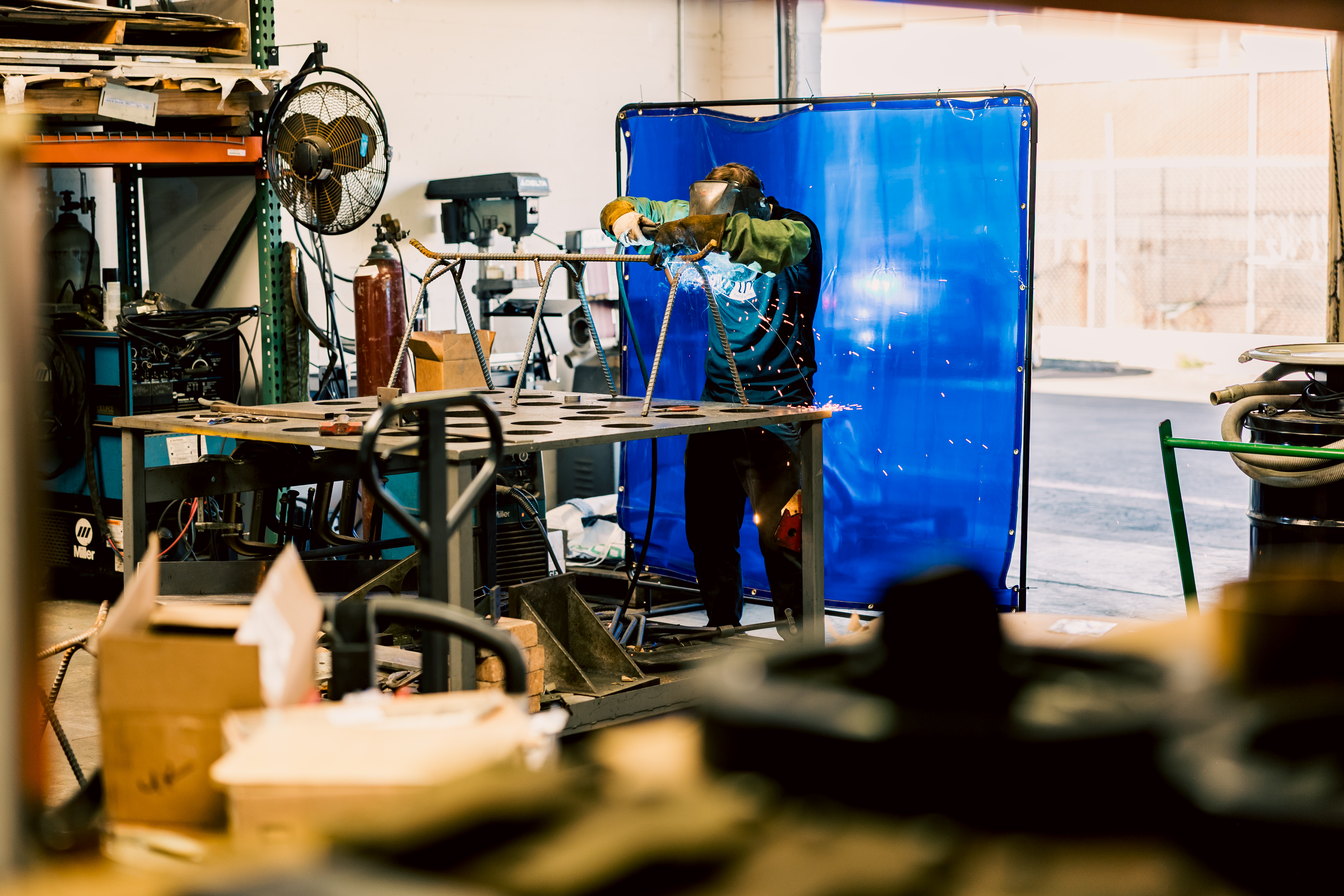 A welder working on custom fabrication behind a blue safety screen in a machine shop.