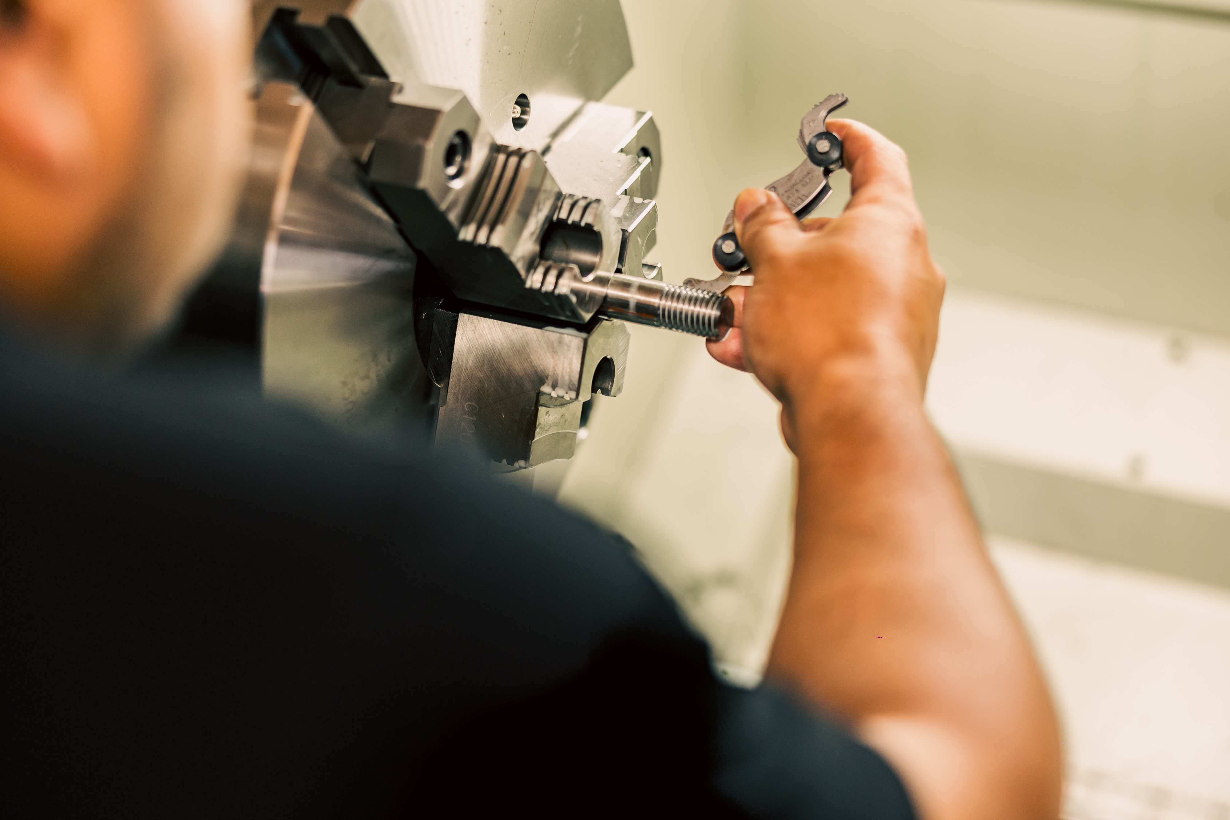 A close-up of a machinist's hands checking the thread pitch on a part using a gauge during a threading operation on a Prototrak 2470.