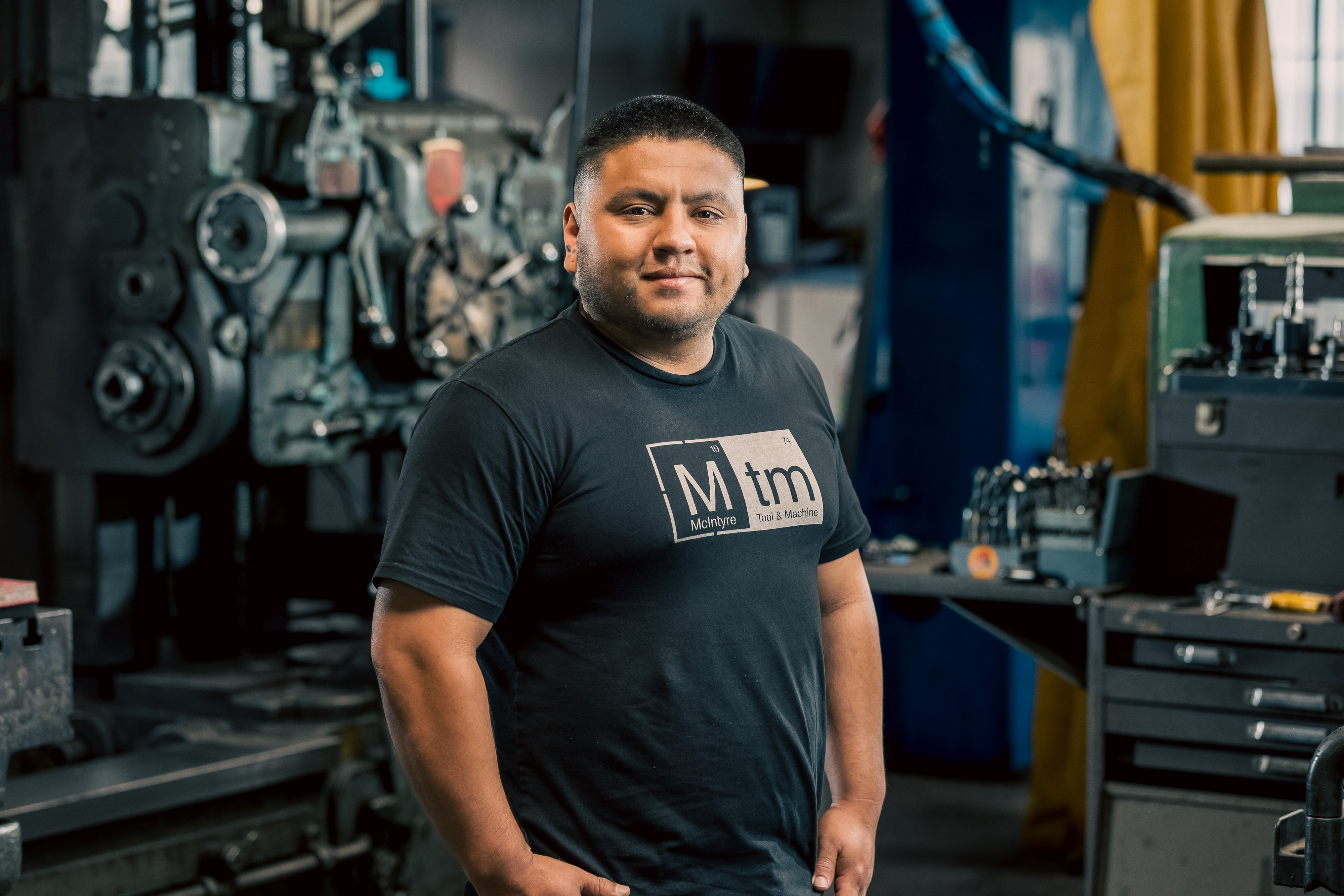 A professional portrait of Carlos, CNC department Lead at MTM, in the machine shop wearing a black MTM shirt.