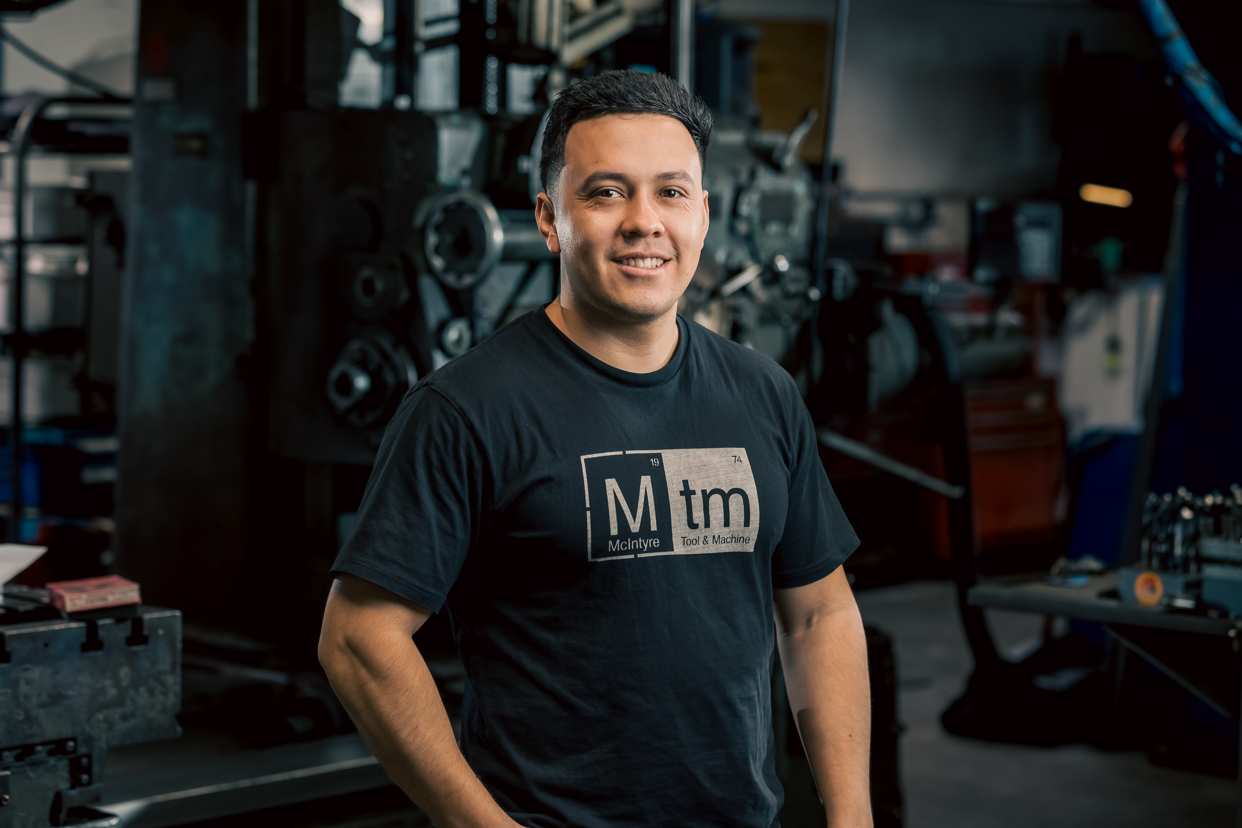 A professional portrait of Miguel, a machine programmer and operator at MTM, smiling in the workshop.