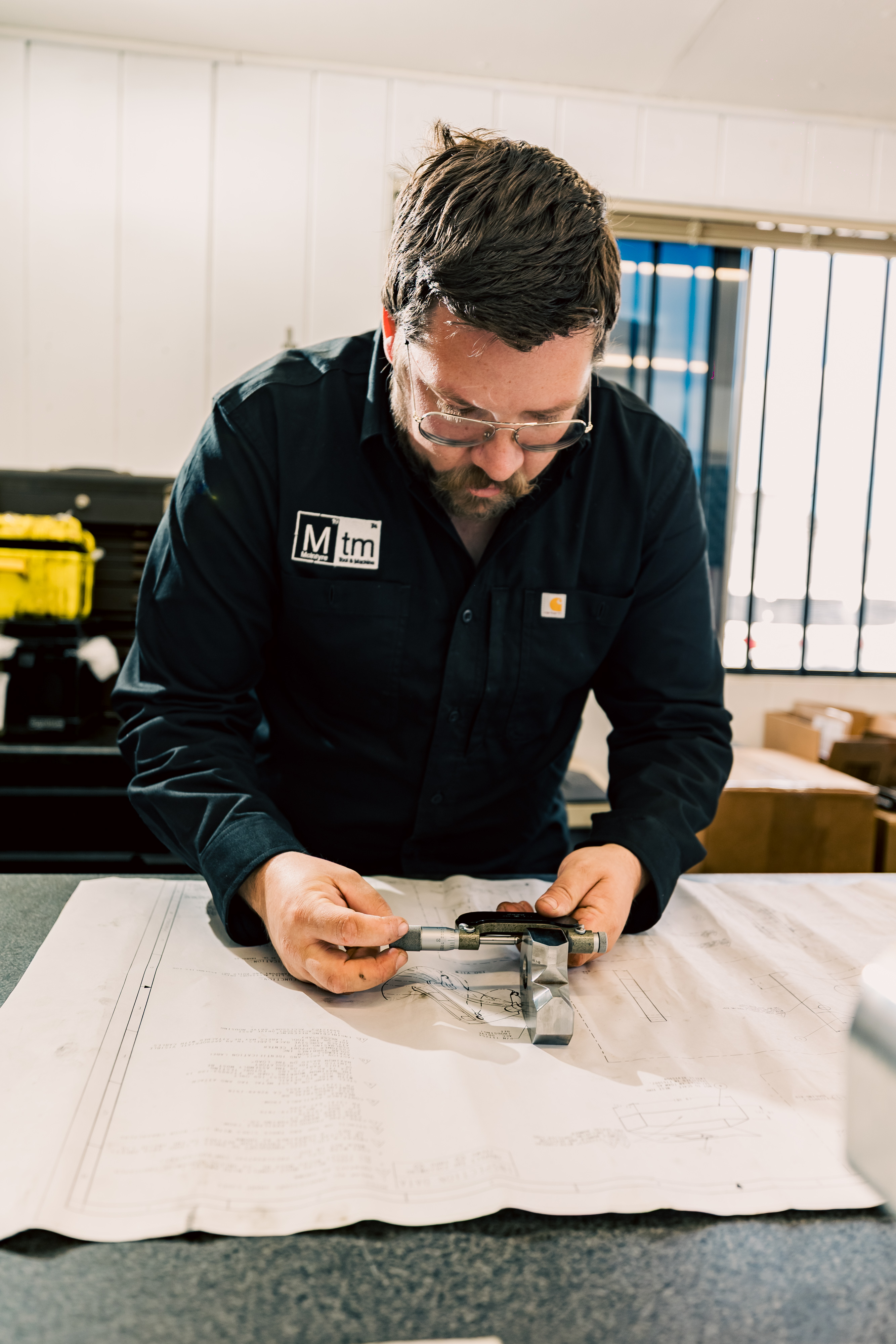 A man in an MTM shirt is performing quality control by verifying part features against a blueprint in the inspection department.
