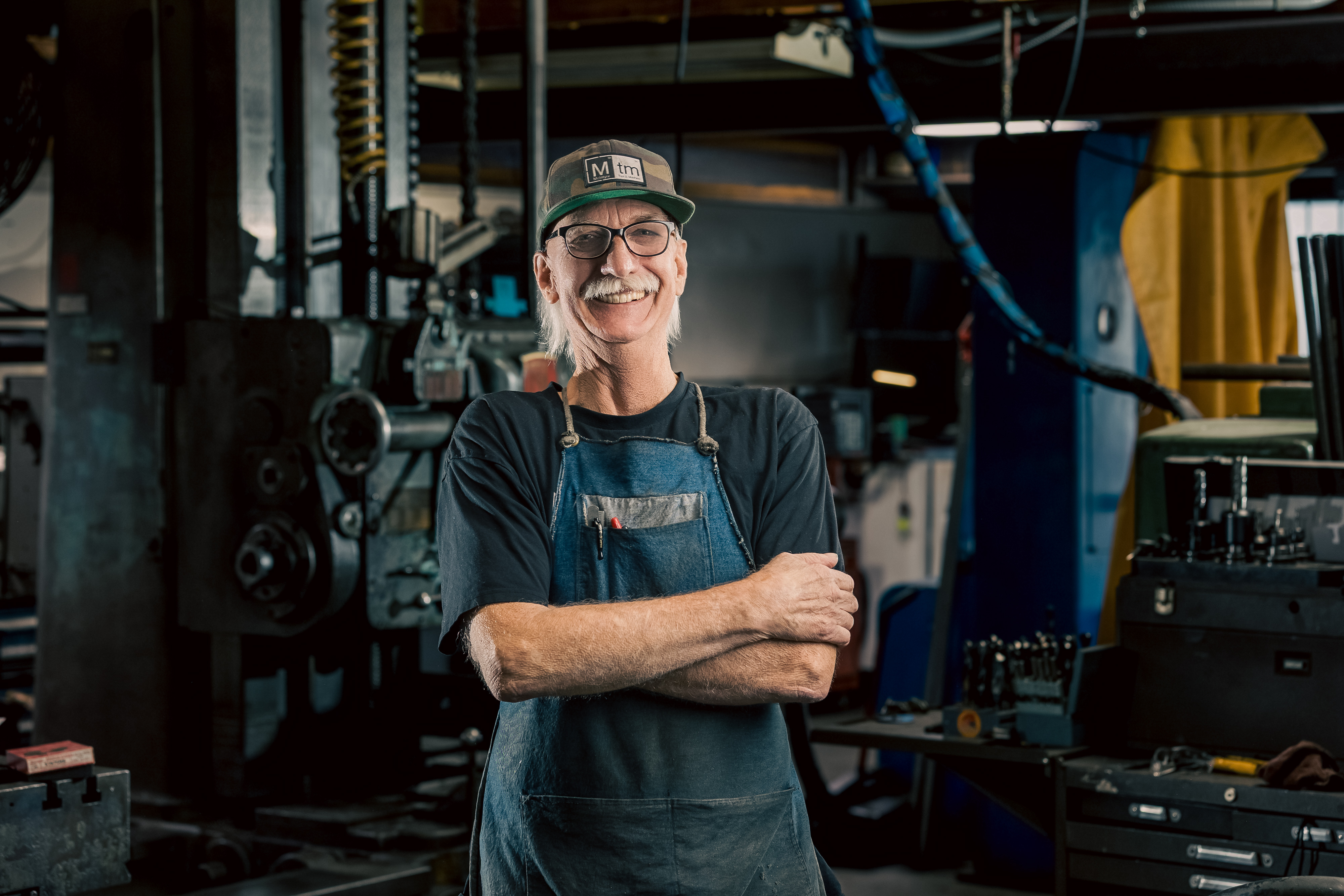 A professional portrait of Ward, Head of the Lathe/turning Department at MTM, wearing a denim apron and cap.