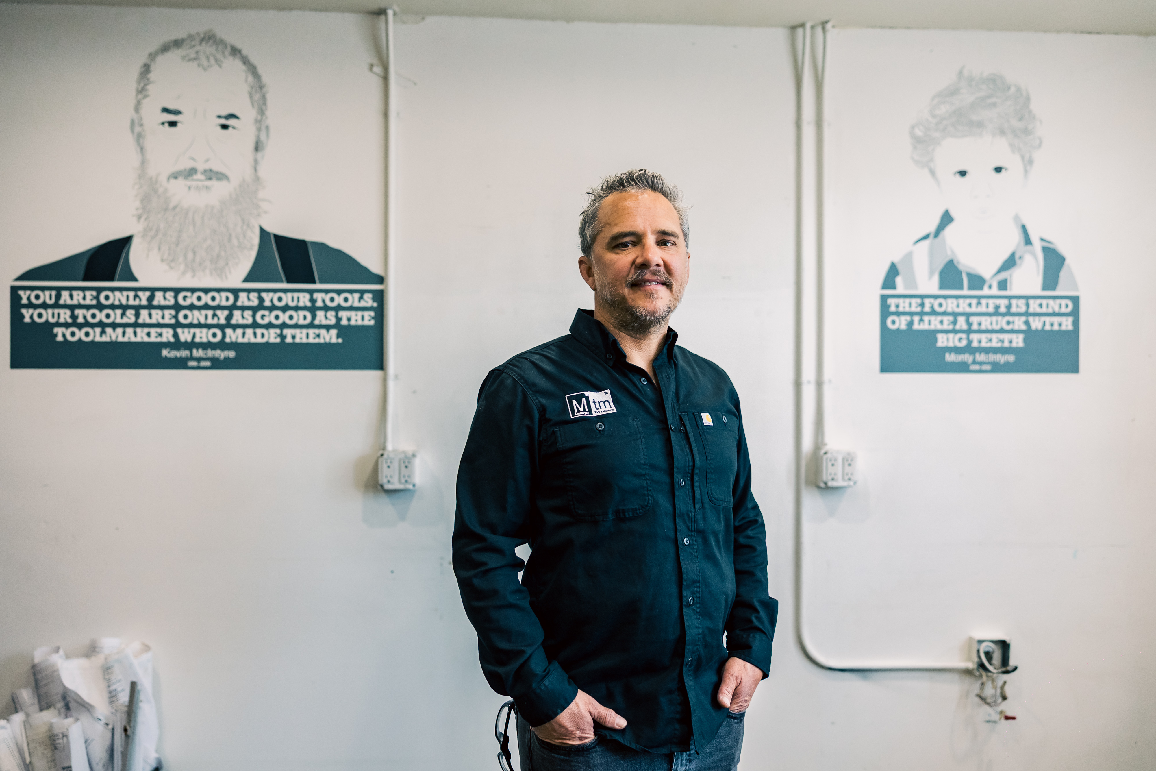 Dustin McIntyre standing between posters of his father and grandfather, representing the legacy of McIntyre Tool & Machine.