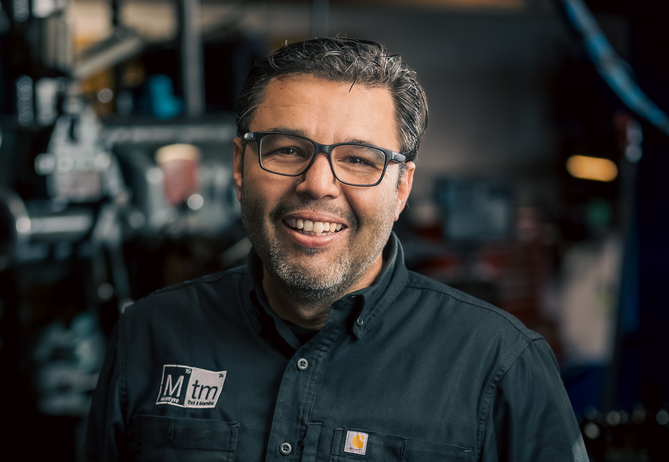 A professional portrait of Pedro Riegas, MTM Operations Manager, smiling in the machine shop wearing a black MTM shirt.
