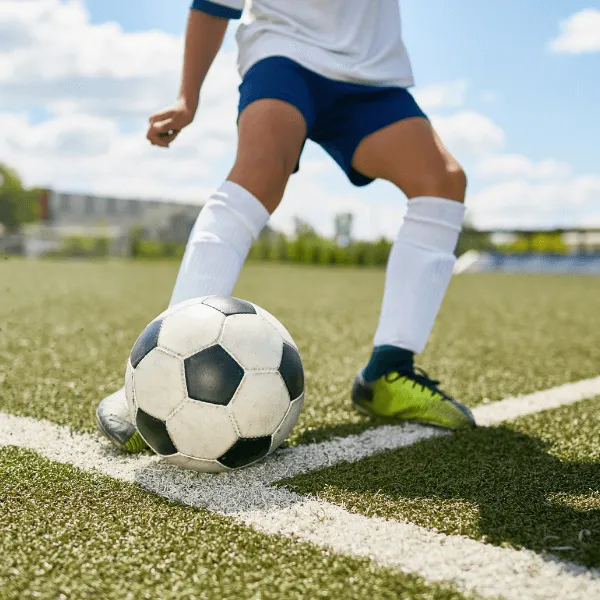 Close-up of a child's legs in a soccer uniform kicking a soccer ball on a green turf field.