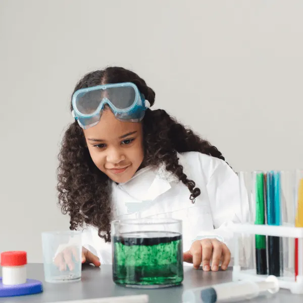 A girl in a white lab coat enthusiastically holding two beakers filled with colorful liquid.
