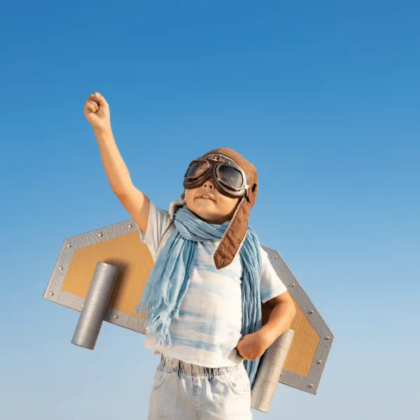 A child running outdoors wearing a handmade cardboard airplane costume and aviator goggles, framed in a circle.