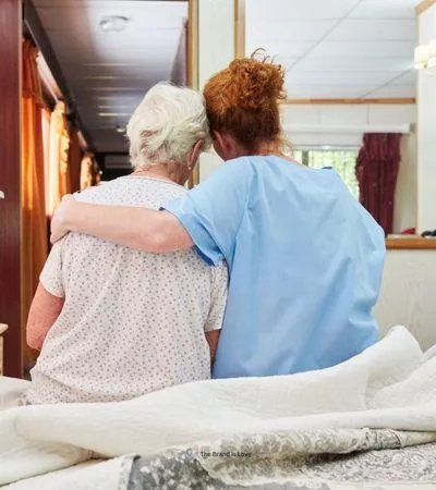 Facility worker with her arm around an elderly woman sitting on a bed, symbolizing hospice and palliative care services coordination.