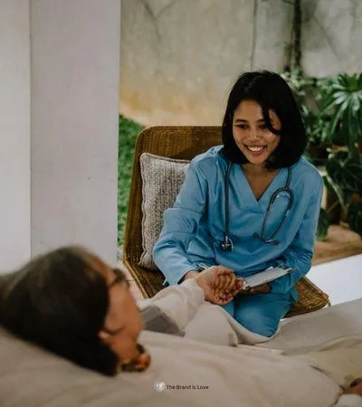 Facility worker sitting in a chair holding the hand of an elderly woman in bed, showcasing how hospice and palliative care coordination lets you focus on your loved one.