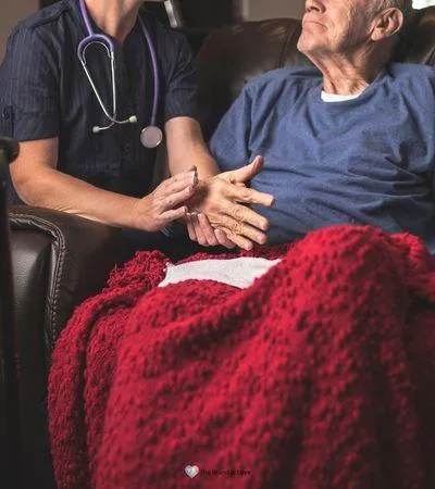 Elderly man sitting in a chair with a red blanket, while a facility care worker kneels next to him, talking with compassion and expertise about hospice care.