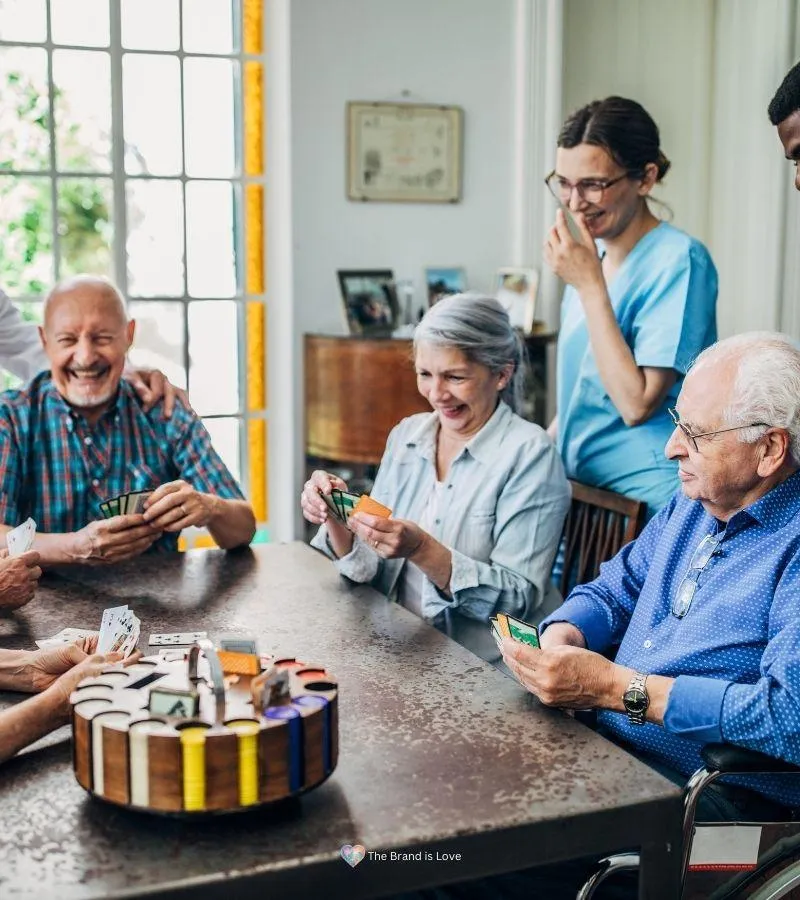 Elder care facility worker standing behind a group of seniors playing a card game, all smiling, symbolizing the possibility of finding the perfect senior living facility.