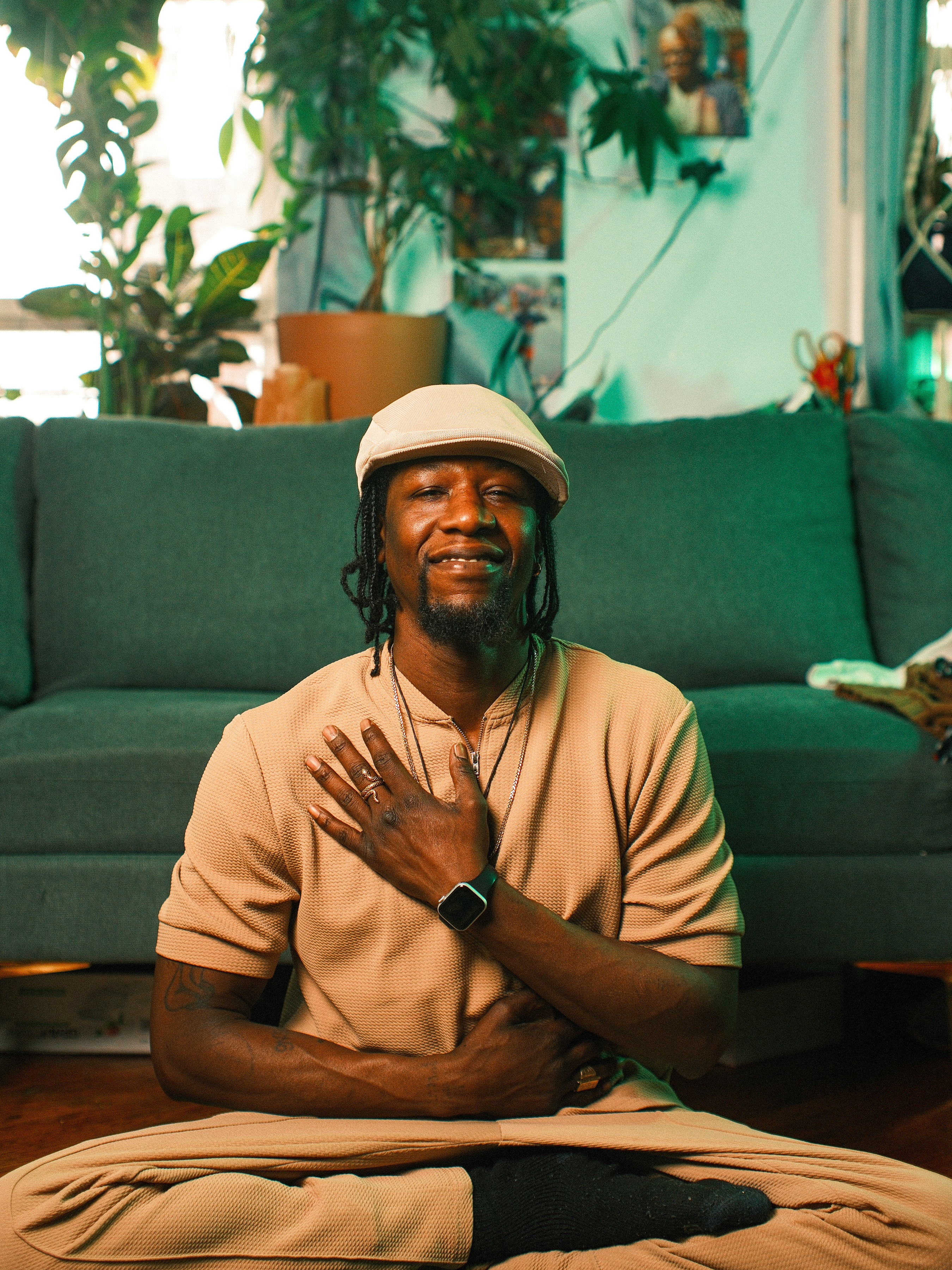 Kwesi Nkroma smiling warmly while sitting cross-legged on the floor, with one hand placed over his heart and the other on his stomach in a centering gesture, set against a backdrop of a teal sofa and lush indoor plants.
