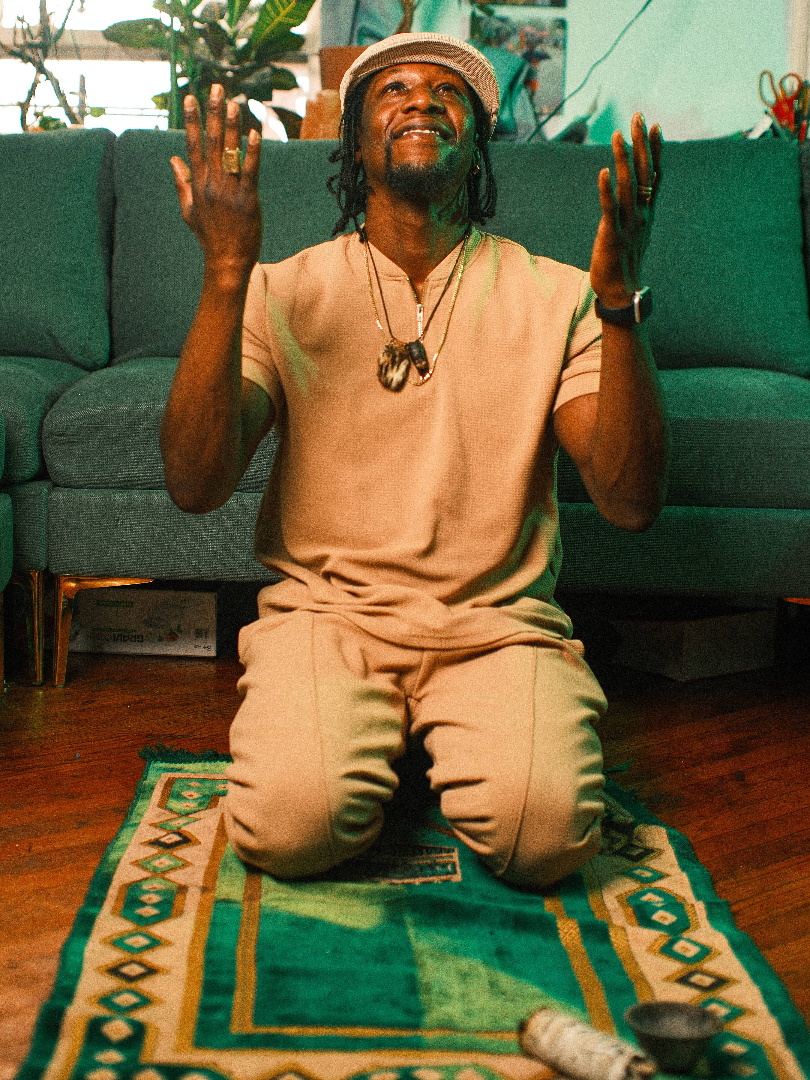 Kwesi Nkroma kneeling on a green patterned prayer rug with his hands raised in a gesture of gratitude or prayer, wearing a tan short-sleeved outfit, a matching cap, and several necklaces in a cozy living room setting.