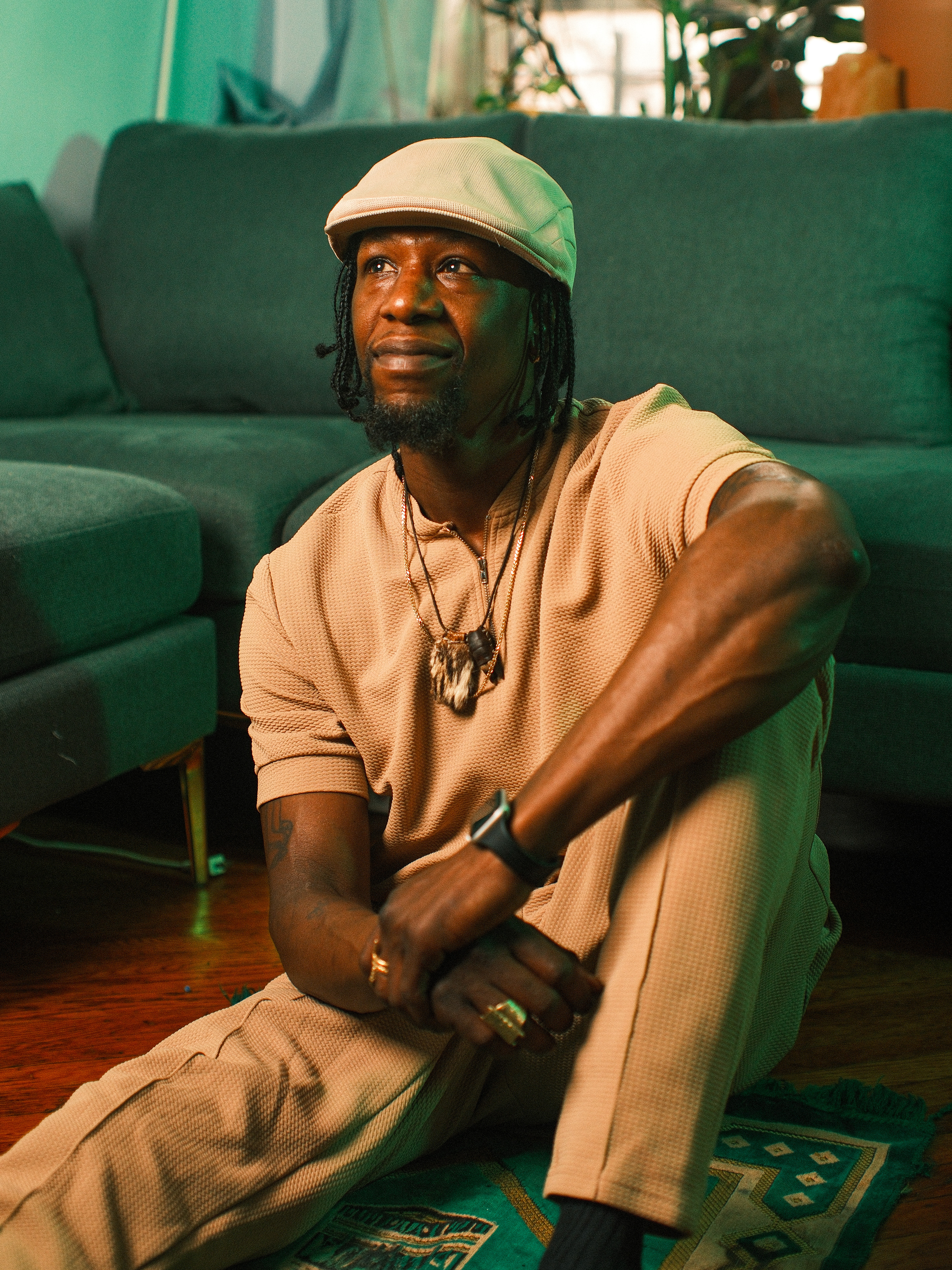 A thoughtful portrait of Kwesi Nkroma sitting on a green prayer rug in a living room, looking upward with a serene expression. He is wearing a textured tan outfit, a flat cap, and layered spiritual necklaces while leaning against a dark teal sofa.