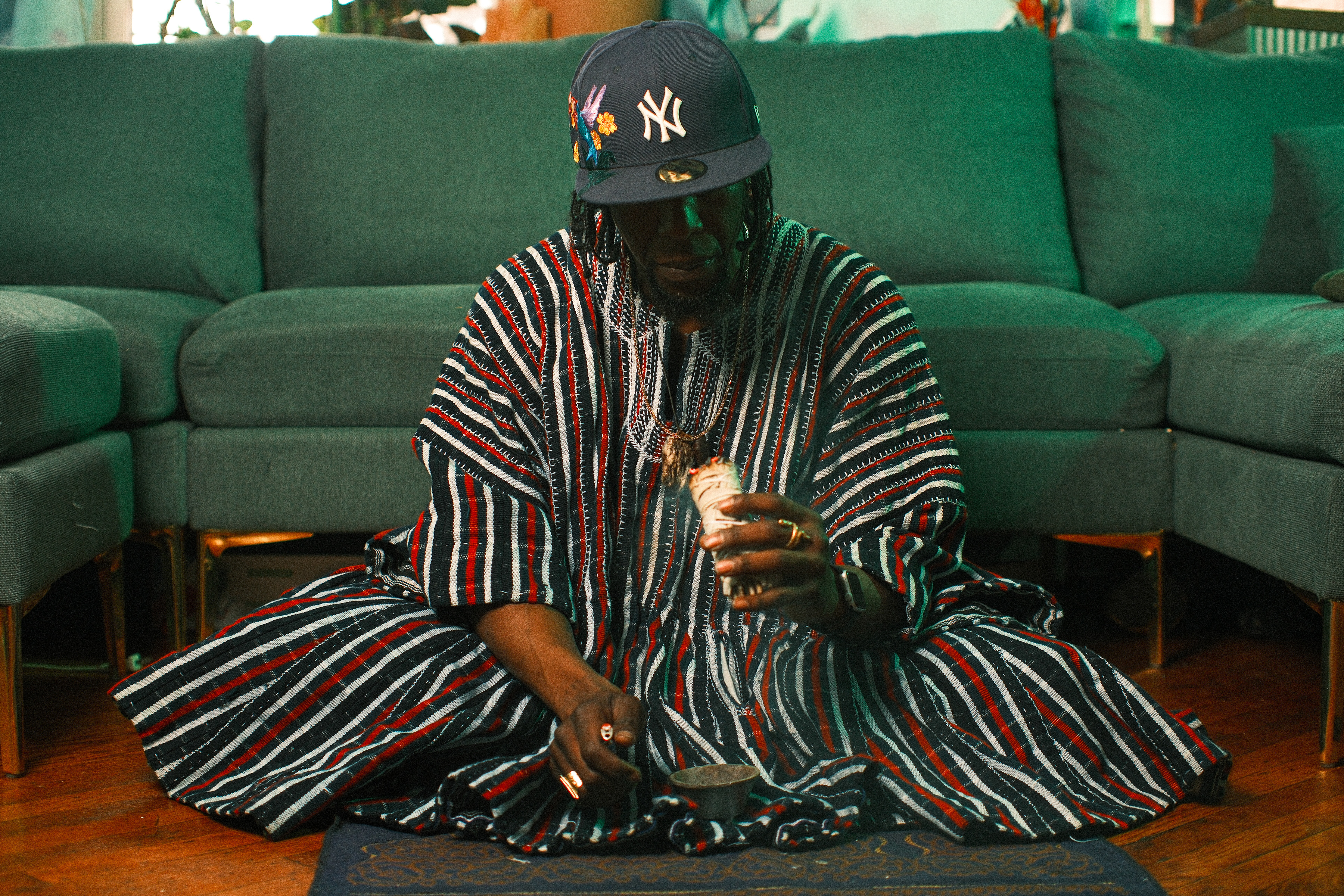 Kwesi Nkroma sitting cross-legged on a rug in a traditional striped West African fugu and a New York Yankees baseball cap, performing a spiritual smudging ceremony with a bundle of burning sage.