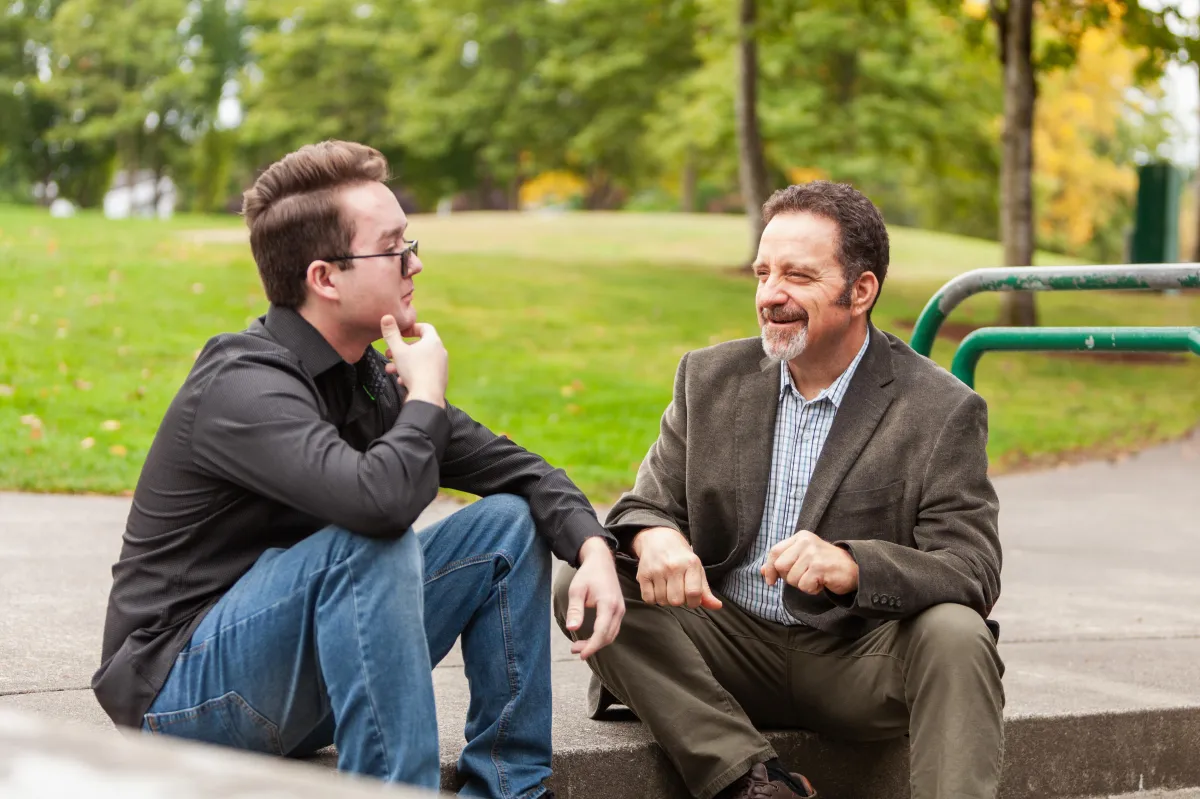 Shannon speaking while John sketches at a sunlit communal table, collaborative scene.