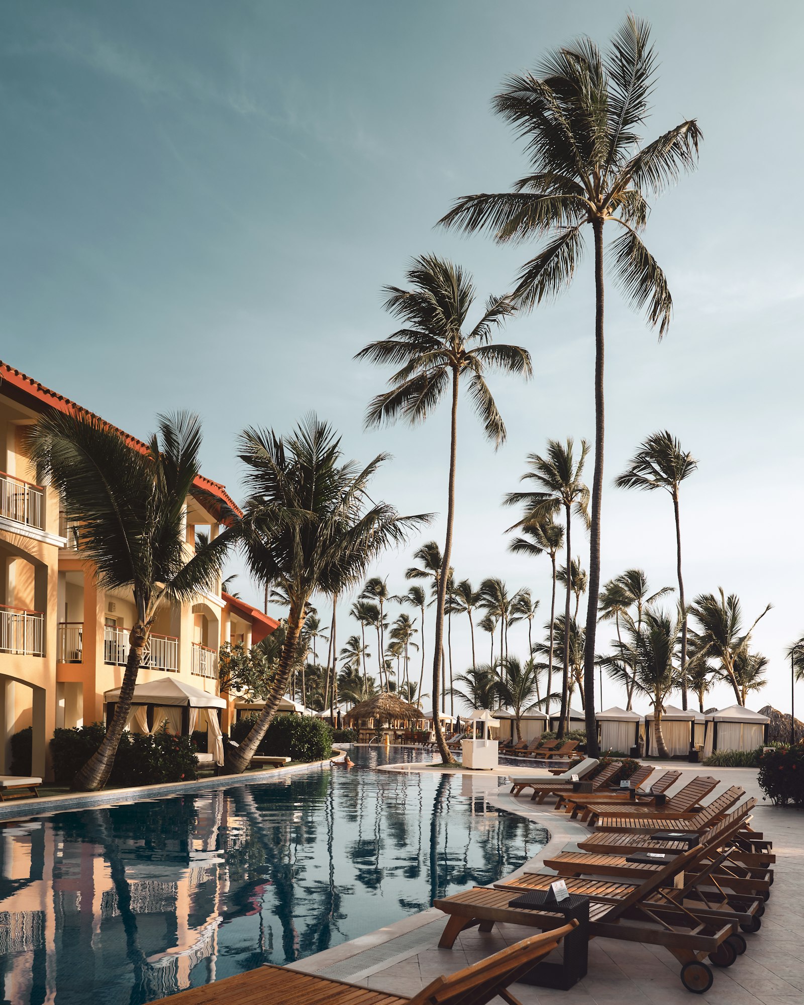 Poolside loungers under tall palm trees at an all-inclusive resort pool at sunset