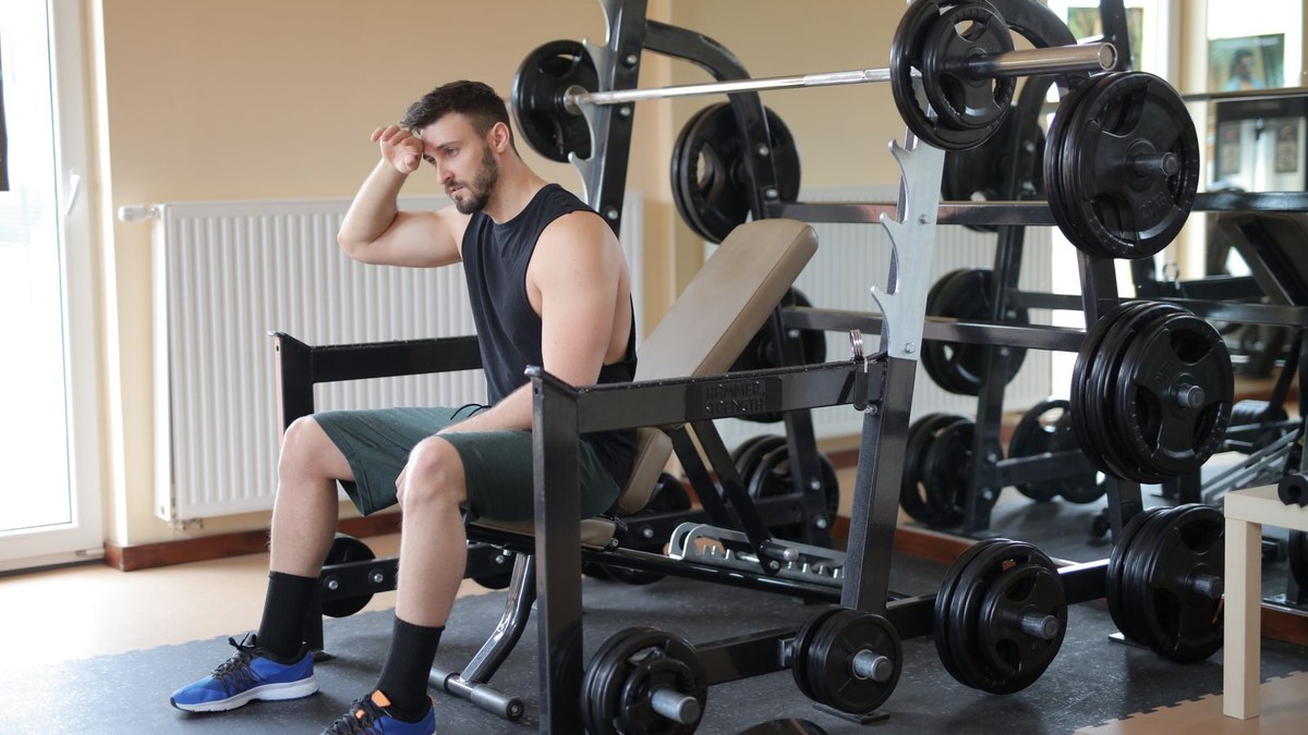 Man sitting on a weight bench in a gym, resting his head on his hand between sets