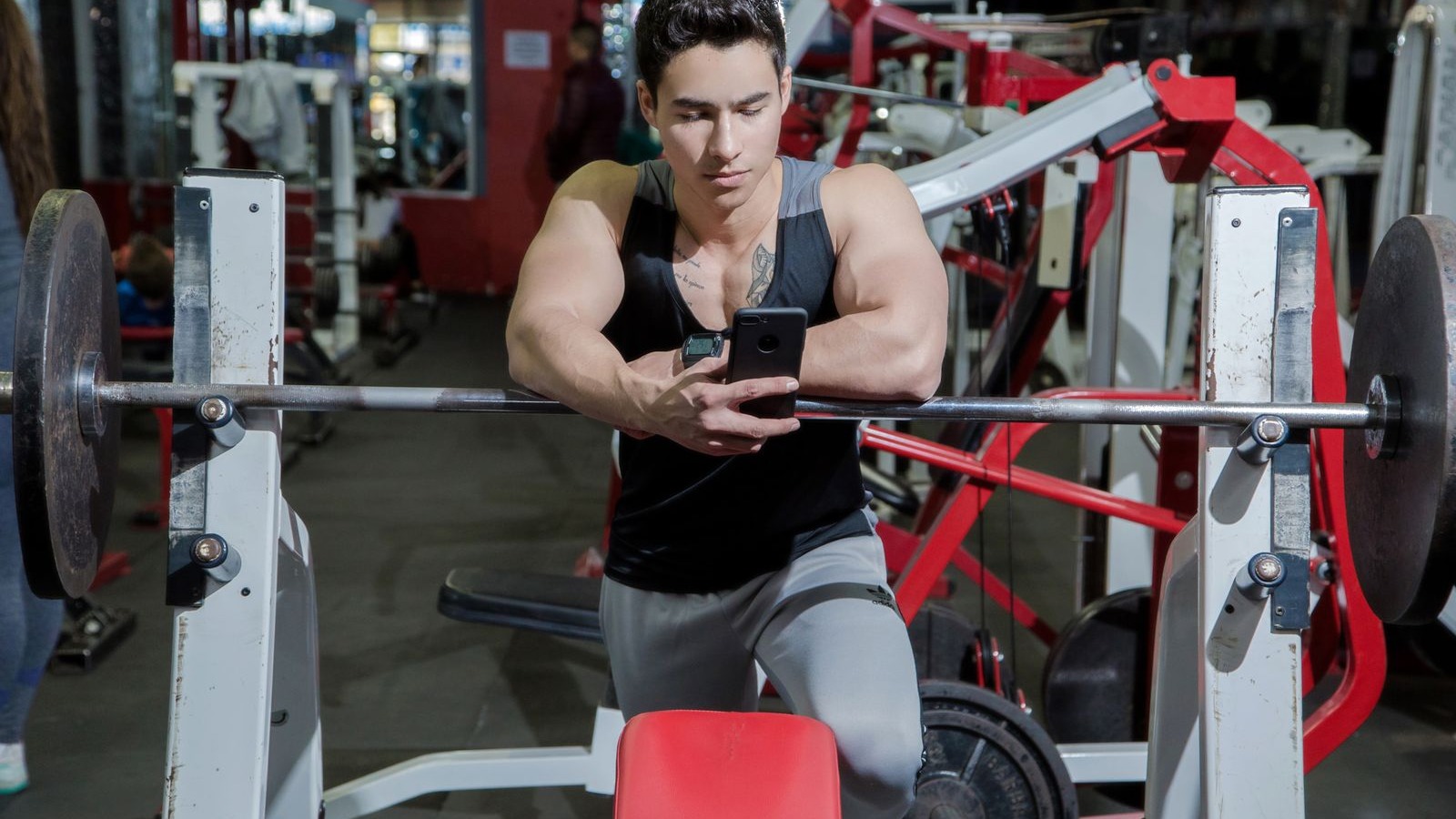 A man sitting on a bench in a gym looking at his phone between sets