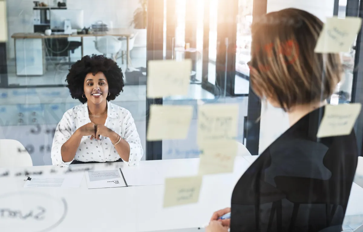 Consultant standing at glass whiteboard with notes facing towards client she is working with, who is seated a a conference table.