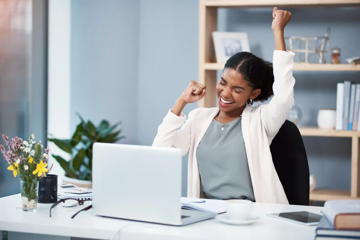 African American Female Business Woman smiling and cheering with hands in the air celebrating while sitting at desk with open laptop and a coffee. She is in an office environment with a window to her left and a bookcase behind her.