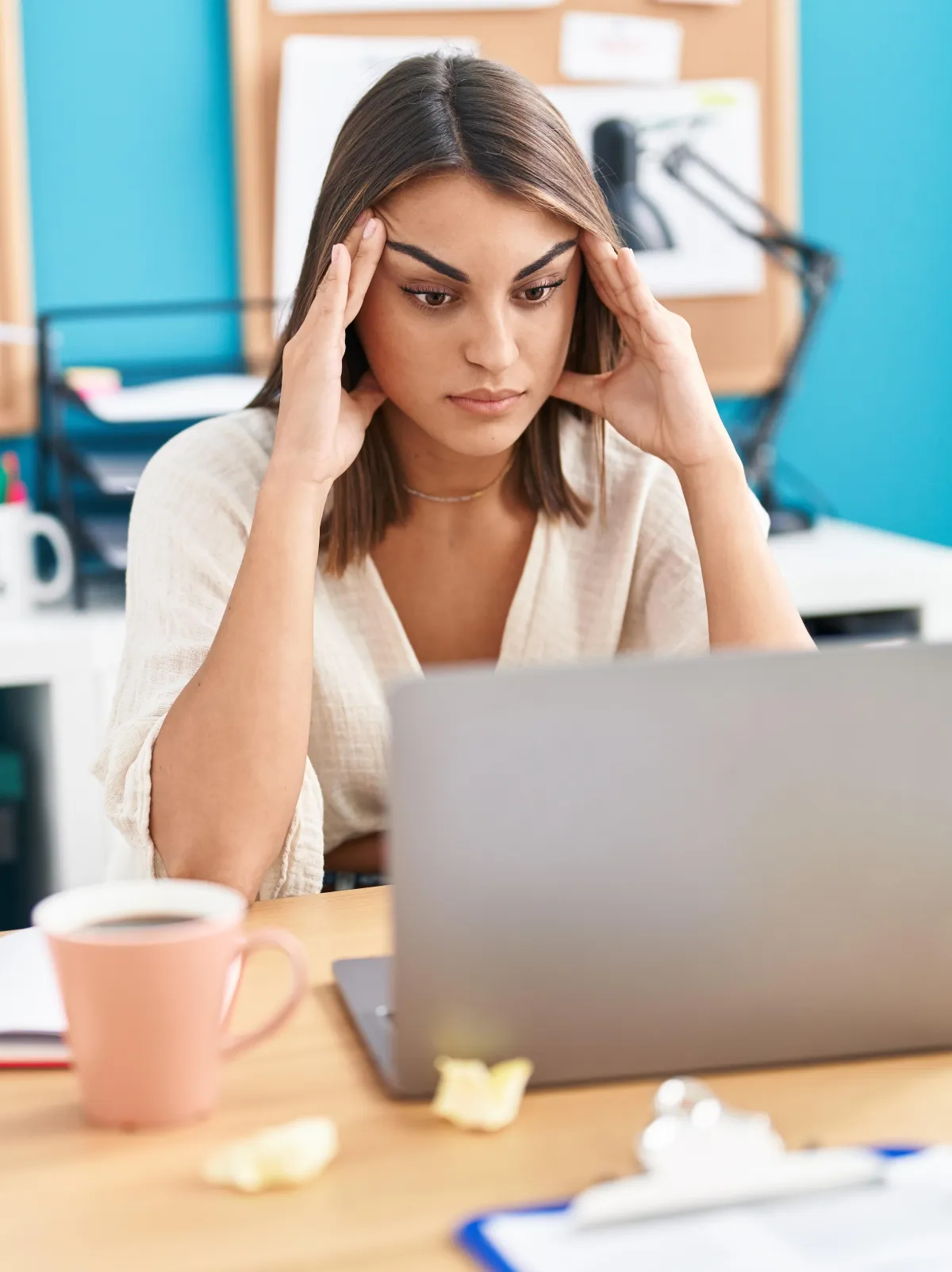 Hispanic business women staring at her laptop, feeling stressed. Her hands are up at her temples. She is sitting at a desk in an office environment.