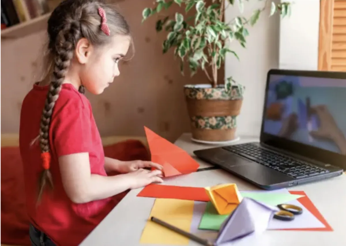 Girl folds paper while watching teacher on laptop