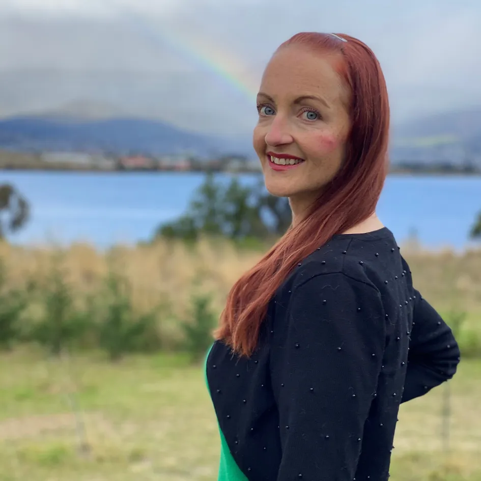 image of Terese Millhouse standing in a field with a river and a rainbow behind her.