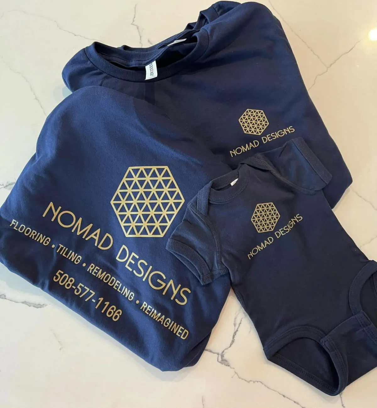 Promotional NOMAD DESIGNS apparel featuring navy blue t-shirts and onesie on a marble background, highlighting flooring, tiling, and remodeling services.