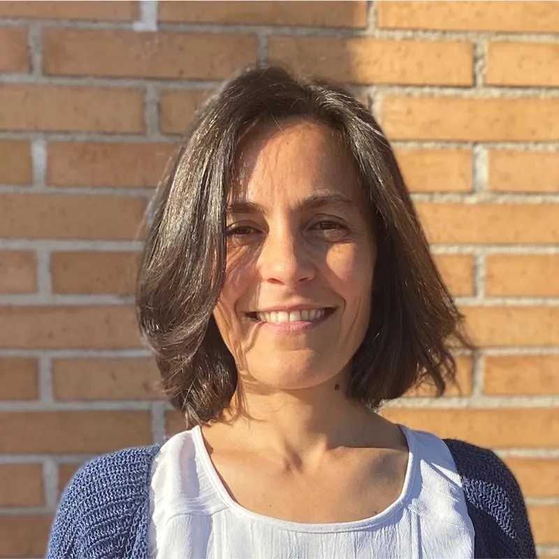 A South Asian woman in her 30s, dressed in a navy blazer, smiling confidently in a modern office with glass walls and greenery. The environment is bright and professional, conveying success and satisfaction.
