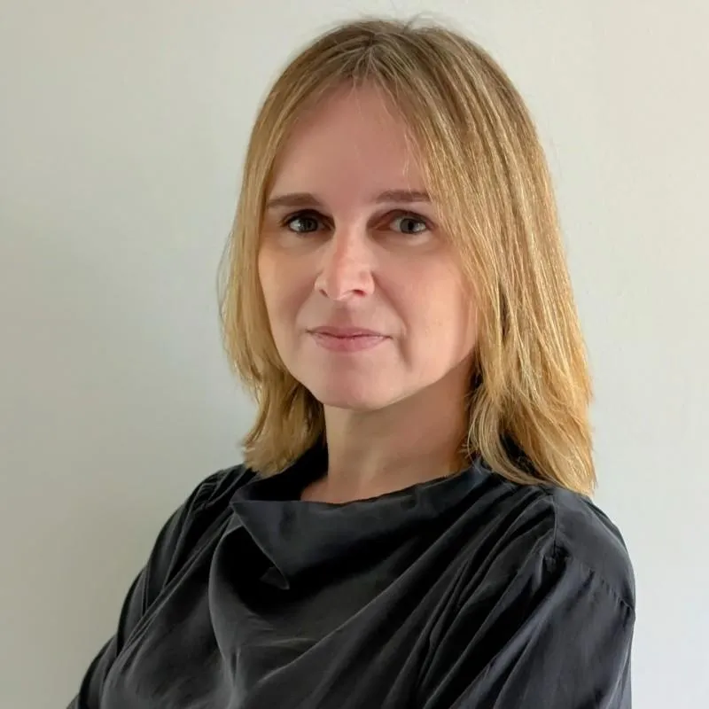A South Asian woman in her 30s, dressed in a navy blazer, smiling confidently in a modern office with glass walls and greenery. The environment is bright and professional, conveying success and satisfaction.