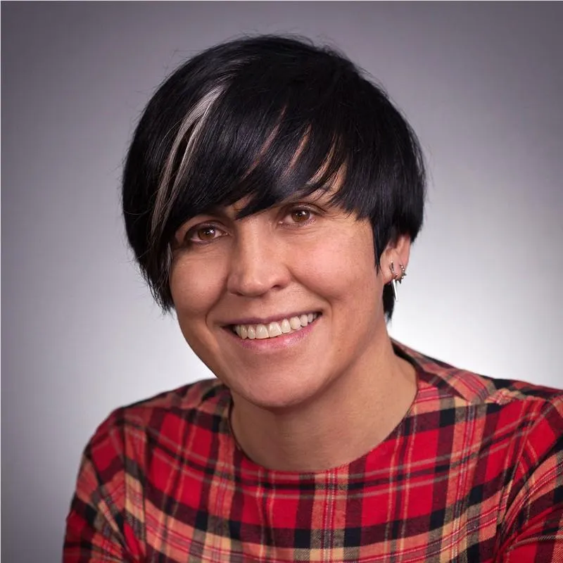 A Black man in his early 40s, wearing a crisp white shirt and glasses, seated at a desk with a laptop and notepad, smiling warmly. The background features bookshelves and soft lighting, evoking trust and achievement.