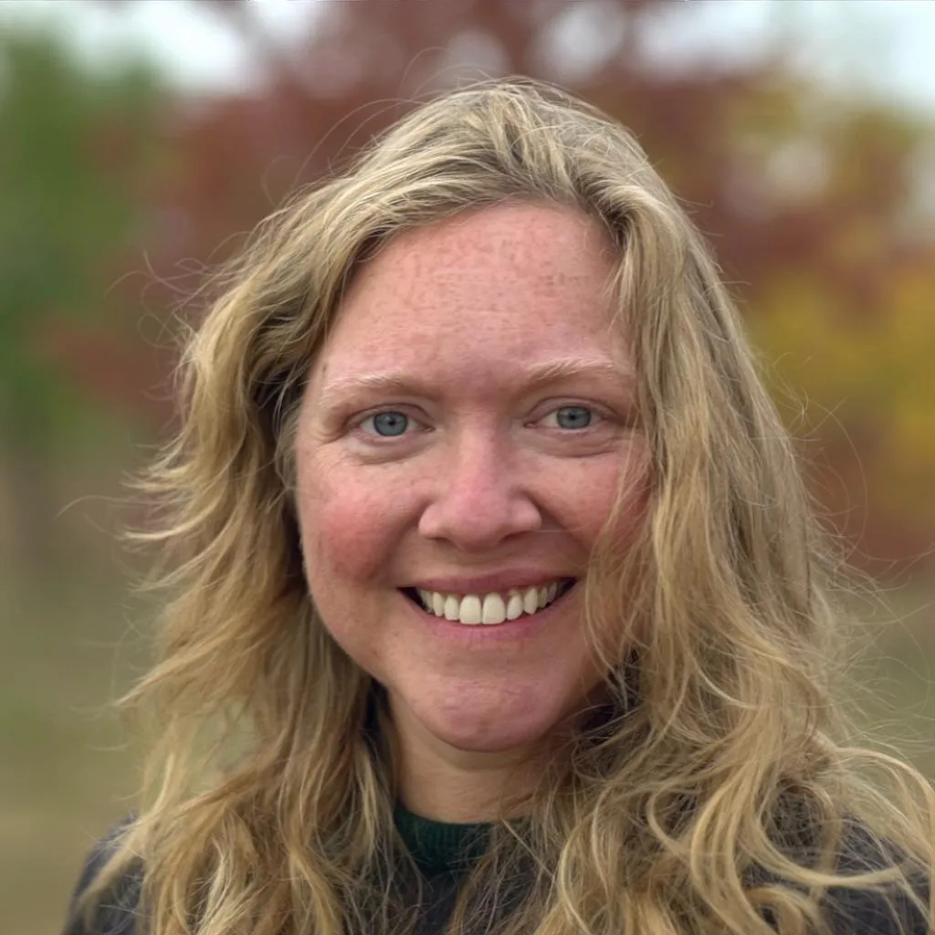 middle-aged woman smiling at the camera, long wavy blonde hair, freckles, blurred trees in he background