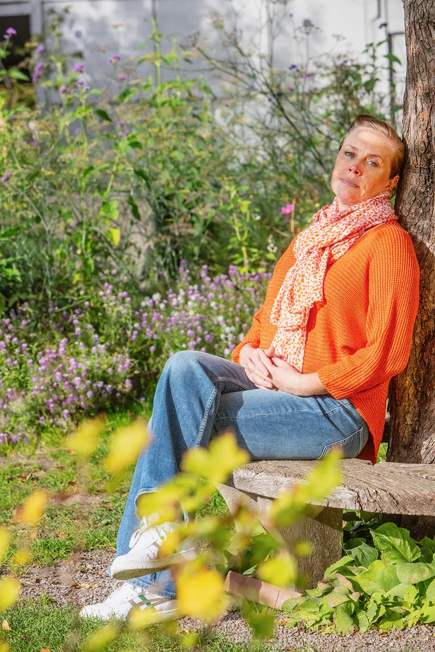 Emmely wearing an orange sweater and patterned scarf, sitting on a wooden bench and leaning against a tree in a garden.